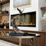 A bronze sculpture of a bird in flight, mounted on a marble base, displayed on a wooden table with a fireplace and various decorative items in the background.